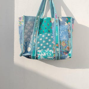 Op de afbeelding: Een patchwork tote bag met verschillende blauwe en turquoise bloemen- en geometrische patronen. De tas heeft stevige handvatten en hangt tegen een witte muur. De randen zijn afgewerkt met een wit en blauw gestreepte stof.