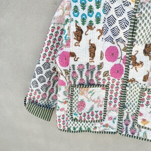 May include: A patchwork jacket with a variety of patterns and colors. The jacket features floral, tiger, and geometric designs in pink, blue, green, and white. The jacket has a green striped trim and two pockets.