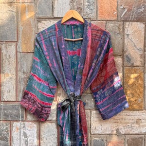 May include: A lightweight kimono robe featuring a multi-colored tie-dye pattern in shades of teal, magenta, and deep blue. The garment has wide sleeves, an open front, and a matching fabric tie belt at the waist, all displayed on a wooden hanger.