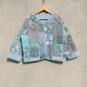 May include: A patchwork jacket with floral patterns in blue, teal, and pink. The jacket has a V-neck and long sleeves with striped cuffs. The jacket is hanging on a wooden hanger.
