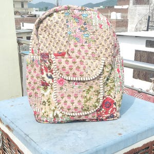 Puede incluir: Una mochila acolchada con un estampado floral en tonos rosa, rojo, verde y beige. La mochila tiene una parte superior redondeada, dos bolsillos laterales y un bolsillo frontal. Ribetes blancos delinean los bolsillos y los bordes.