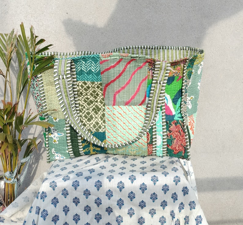 May include: A patchwork tote bag with a green, blue, and pink color scheme. The bag has a large, rectangular shape and two handles. The bag is made of fabric and has a quilted design.