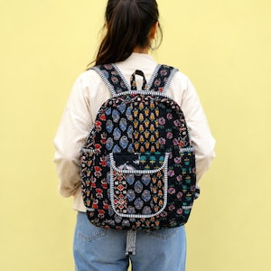 Puede incluir: Una mochila acolchada con base negra y un patrón de estampado de bloques floral y geométrico multicolor. La bolsa incluye un bolsillo frontal con solapa, correas ajustables para los hombros y ribetes a rayas blancas y negras.
