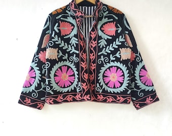 Puede incluir: Chaqueta negra bordada con motivos florales coloridos. La chaqueta presenta diseños florales rosas, celeste y naranjas. Cuelga de una percha de madera contra una pared blanca.