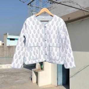 以下が含まれることがあります： 白と青の花柄キルティングジャケット。丸襟、長袖、フロントポケット2つ付き。木製のハンガーに掛けられ、ボタンで留められています。画像には寸法は表示されていません。