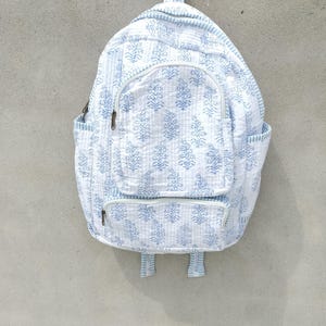 Puede incluir: Una mochila acolchada con base blanca y un patrón repetitivo de estampado floral en bloque de color azul claro. La bolsa incluye un compartimento principal grande, un bolsillo frontal con cremallera, dos bolsillos laterales, un asa superior y correas ajustables.