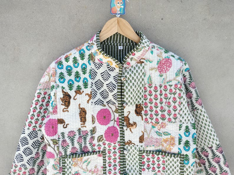 May include: A quilted jacket with a patchwork design. The jacket features various patterns, including floral, geometric, and animal prints in shades of pink, blue, green, and brown on a white background. The jacket is hanging on a wooden hanger.