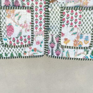 May include: Close-up of a quilted jacket with a floral pattern in pink, green, and yellow on a white background. The jacket has green striped cuffs and pocket trim. The design includes a mix of floral and geometric patterns.