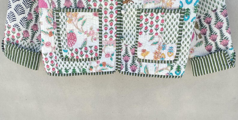 May include: Close-up of a quilted jacket with a floral pattern in pink, green, and yellow on a white background. The jacket has green striped cuffs and pocket trim. The design includes a mix of floral and geometric patterns.