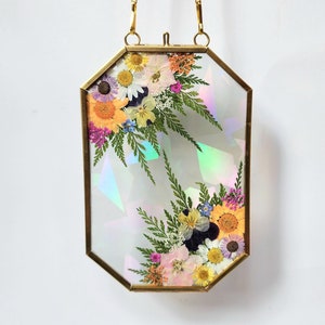 May include: A gold octagonal frame with a chain for hanging, featuring pressed flowers and iridescent foil paper. The flowers are arranged in two bouquets, one on each side of the frame.