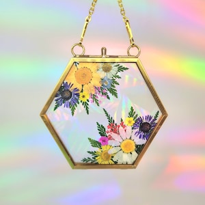 May include: A hexagonal gold frame with a chain for hanging, containing a pressed flower arrangement with a variety of colors, including yellow, white, purple, and pink.