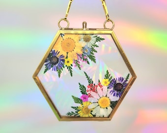 Può includere: Una cornice esagonale dorata con una catena per appenderla, contenente una disposizione di fiori pressati con una varietà di colori, tra cui giallo, bianco, viola e rosa.