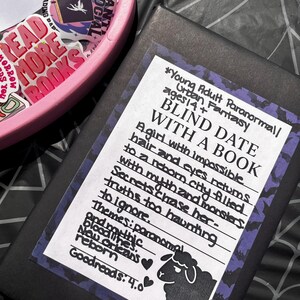 May include: A wrapped black book with a white label detailing a young adult paranormal novel, titled "Blind Date with a Book." A pink container filled with stickers is visible in the background.
