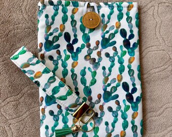 May include: A white fabric pouch with a green cactus pattern, a matching wrist strap, and a gold-colored keychain with a green tassel. The pouch has a wooden button closure.
