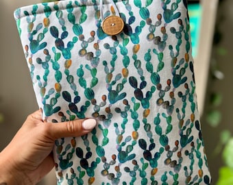 May include: A white book sleeve featuring a repeating pattern of green and blue cacti with orange accents. The sleeve is secured with a white elastic loop and a wooden button closure. The book sleeve is held in a person's hand.