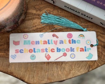 Pode incluir: Um marcador de página branco com um design colorido e o texto "Mentally at the scholastic book fair". O marcador de página tem uma borla turquesa.