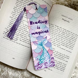 Puede incluir: Un marcapáginas morado y rosa con una cola de sirena y el texto "Reading is magical".