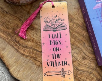 May include: A pink and orange bookmark with a book, stars, and a sword. The bookmark has the text "I call dibs on the villain."