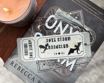 May include: A book titled "One Dark Window" by Rebecca Yarros, a #1 New York Times bestselling author. The book cover features a silver bookmark with a dragon design and the text "Onyx Storm Bookclub".