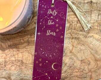May include: A burgundy bookmark with a gold tassel. The bookmark features a gold design of constellations and the text "Defy the Stars".