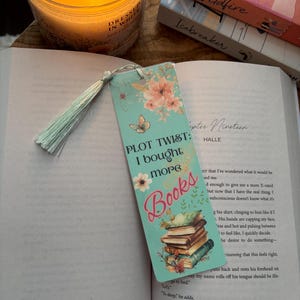 May include: A teal blue bookmark featuring a stack of books, flowers, and a butterfly. The bookmark has the text "PLOT TWIST: I bought more BOOKS" on it.