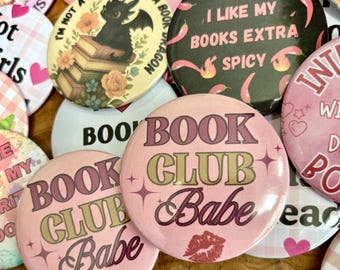 May include: A collection of round, colorful buttons with various designs and text. Some buttons feature phrases like "Book Club Babe" and "I like my books extra spicy." Other designs include a dragon on books and floral accents. The buttons are in shades of pink, black, and white.