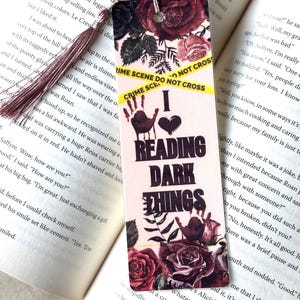 May include: A bookmark with a floral design featuring burgundy roses and the text "I Love Reading Dark Things." The bookmark has a tassel and yellow "Crime Scene Do Not Cross" tape.