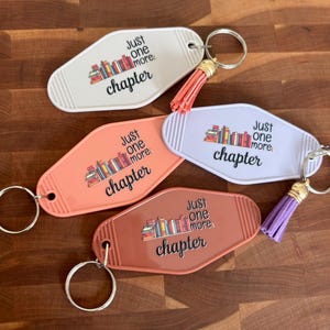 Puede incluir: Cuatro llaveros estilo motel en blanco, melocotón, lila y marrón. Cada llavero tiene una colorida ilustración de libros y el texto "Just one more chapter". Los llaveros tienen anillos metálicos y borlas.