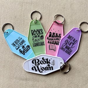 May include: A selection of colourful keyrings with book-related quotes. The keyrings are in shades of blue, green, pink, purple, and white. Each keyring features a different quote, including "The book is always better" and "Book Worm".