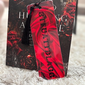 May include: A red bookmark with a black tassel and the word "SITUATIONAL" printed in black on it. The bookmark is laying on a white fuzzy surface.