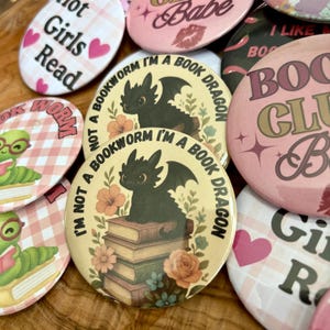 Peut inclure: Une collection de badges ronds et ovales avec divers motifs et textes. Certains badges affichent des phrases telles que "Hot Girls Read" et "Book Club Babe". Un badge présente une illustration de dragon noir avec le texte "I'm Not a Bookworm I'm a Book Dragon."
