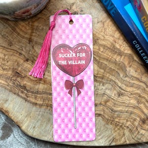 May include: Pink bookmark with a pink and white quilted pattern. A red heart-shaped lollipop with a white stick and a red bow is on the bookmark. The lollipop says "SUCKER FOR THE VILLAIN".