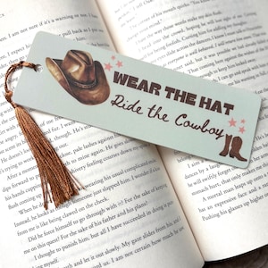 May include: A light blue bookmark featuring an illustration of a cowboy hat and boots. The bookmark has the text "Wear the Hat Ride the Cowboy" in a vintage style font.