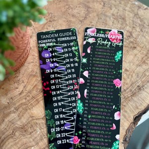以下が含まれることがあります： 白文字の黒いブックマークが2つあります。1つは「Tandem Guide Powerful Powerless」と章リストが書かれています。もう1つは「Fearless/Fearful Reading Guide」と章リストが書かれています。どちらにも装飾的な花と宝石のアクセントがあります。