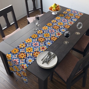 May include: A blue, yellow, and red table runner with a geometric tile pattern. The runner is on a dark wood table with two chairs and a bowl of fruit.