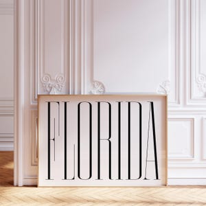 Op de afbeelding: Een ingelijste kunstdruk met het woord "FLORIDA" in een vetgedrukte, zwarte letter met schreef. De print staat tegen een witte achtergrond en is omlijst in licht hout. Het kunstwerk wordt tentoongesteld op een houten vloer, met witte lambrisering op de achtergrond.