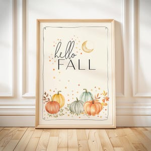 May include: A framed art print with a light beige background. The print features the words "hello FALL" in black script, a crescent moon, and watercolor pumpkins in orange, yellow, and green. The pumpkins are surrounded by leaves and stars.