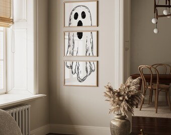 May include: Three framed black and white ghost art prints hang on a beige wall. The ghost illustrations have a simple, cartoonish style. A black bat garland hangs above the frames, and a pumpkin sits on a table in front of the wall.
