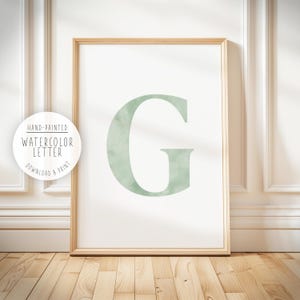 Puede incluir: Un cuadro enmarcado con la letra "G" en acuarela, en un tono verde suave. La obra de arte está sobre un fondo blanco y enmarcada en madera clara. Un gráfico circular dice "Hand-Painted Watercolor Letter Download & Print."