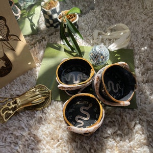 Puede incluir: Tres posavasos de cerámica negra con ribete dorado y un diseño de serpiente blanca. Cada posavasos presenta un símbolo del zodíaco diferente.