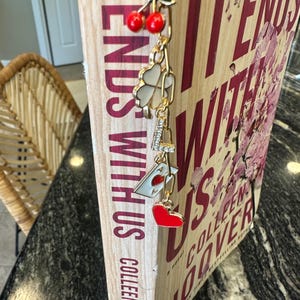 May include: A gold bookmark with charms, including a four-leaf clover, a heart, a cherry, and the letter "L". The bookmark is attached to a book titled "It Ends With Us" by Colleen Hoover.