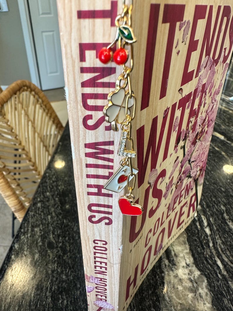 May include: A gold bookmark with charms, including a four-leaf clover, a heart, a cherry, and the letter "L". The bookmark is attached to a book titled "It Ends With Us" by Colleen Hoover.