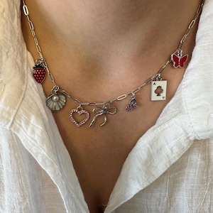 May include: A silver chain necklace with various charms, including a strawberry, a seashell, a heart, a bow, a cherry, a playing card, and a butterfly. The charms are all silver and have red, pink, and white enamel accents.