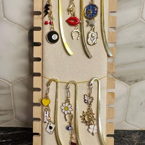 May include: A set of six gold bookmark holders with various charms hanging from them. The charms include a black 8 ball, a red lip, a horseshoe, a white flower, a yellow heart, a daisy, a butterfly, a sun, a four-leaf clover, a playing card, an evil eye, and a wine glass.