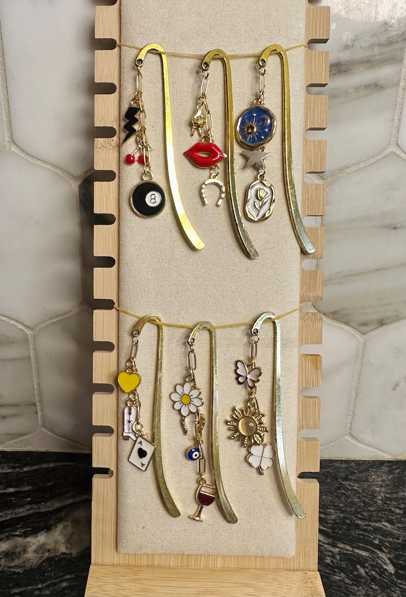 May include: A set of six gold bookmark holders with various charms hanging from them. The charms include a black 8 ball, a red lip, a horseshoe, a white flower, a yellow heart, a daisy, a butterfly, a sun, a four-leaf clover, a playing card, an evil eye, and a wine glass.