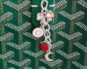 May include: A green Goyard bag with a silver charm attached to the zipper pull. The charm features a bow, a letter C, a strawberry, and a crescent moon.