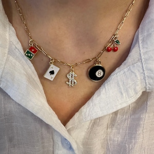 May include: A gold chain necklace with multiple charms, including a playing card, a dollar sign, a cherry, and a black and white 8-ball.