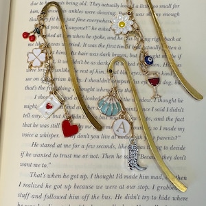 May include: A gold bookmark with a variety of charms, including a red heart, a white four-leaf clover, a white horseshoe, a blue and white umbrella, a red wine glass, a blue eye, and a silver cowboy boot. The bookmark is resting on a page of a book with text about a boy and his friend.