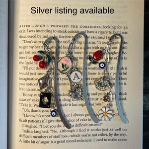 May include: Silver bookmark with charms, including a red cherry, a heart, a letter A, a snowflake, a lips charm, a daisy, and a blue evil eye. The bookmark is made of metal and has a silver finish. The bookmark is on a book page with text that reads "AFTER LUNCH I PROWLED THE CORRIDORS, looking for an exit. I was intending to sneak outside and have a cigarette, but I was discovered by Indira by the fire escape. He assumed I was..."