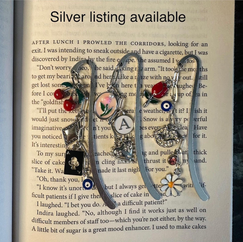 May include: Silver bookmark with charms, including a red cherry, a heart, a letter A, a snowflake, a lips charm, a daisy, and a blue evil eye. The bookmark is made of metal and has a silver finish. The bookmark is on a book page with text that reads "AFTER LUNCH I PROWLED THE CORRIDORS, looking for an exit. I was intending to sneak outside and have a cigarette, but I was discovered by Indira by the fire escape. He assumed I was..."